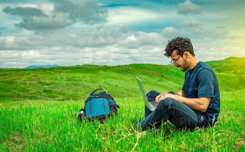 Smiling freelancer young man with laptop working from the field. Close up of man sitting on green grass working with his laptop. Smiling man with laptop sitting on the grass in the field