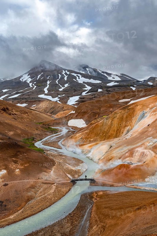Tourist on bridge over steaming stream between colourful rhyolite mountains with snowfields, Hveradalir geothermal area, Kerlingarfjöll, Icelandic highlands, Iceland, Europe