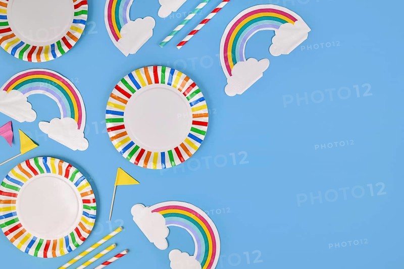 Party flat lay with colorful plates, rainbow napkins and drinking straws on blue background with copy space