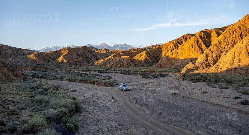 Off-road vehicle driving through a canyon, landscape of eroded hills at sunrise, badlands, white mountain peaks of the Tian Shan Mountains in the background, Canyon of the Forgotten Rivers, Issyk Kul, Kyrgyzstan, Asia