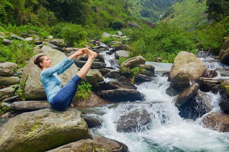 Yoga exercise outdoors, panorama of woman doing Ashtanga Vinyasa Yoga balance asana Ubhaya padangusthasana Big Double Toe Yoga Pose at tropical waterfall in Himalayas in India. Panoramic image