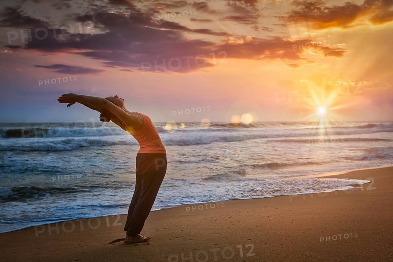 Young sporty fit woman doing yoga Sun salutation Surya Namaskar pose Hasta Uttanasana on tropical beach on sunset. With lens flare