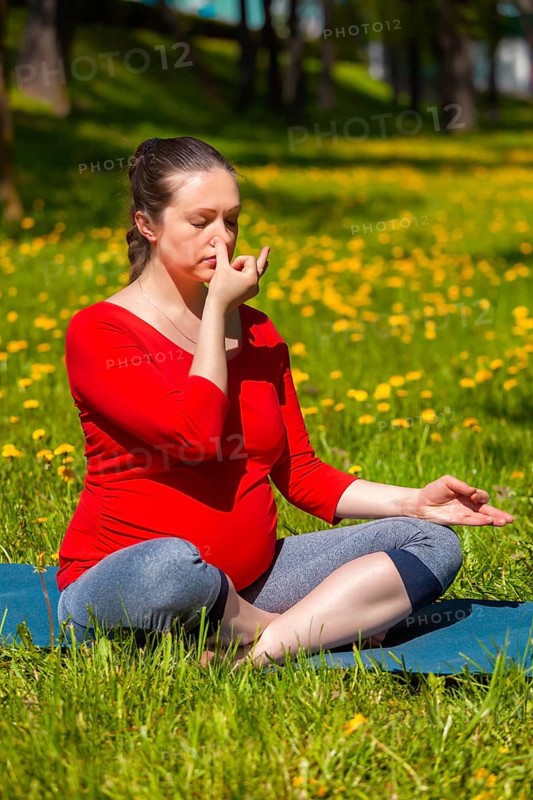 Pregnancy yoga exercise, pregnant woman doing pranayama breath exercise in Sukhasana easy yoga pose with crossed legs outdoors on grass lawn with dandelions in summer