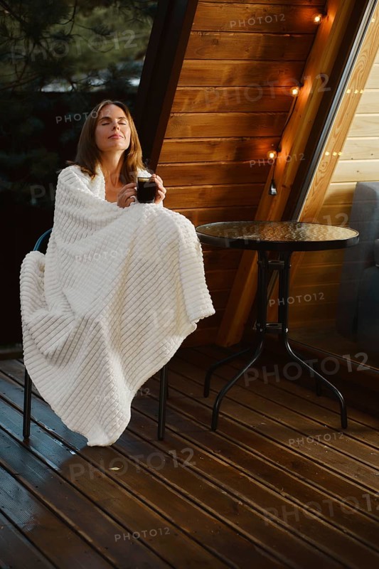 Dreamy young woman sits on terrace of a log cabin wrapped in a blanket with a cup of herbal tea and enjoying the fresh air