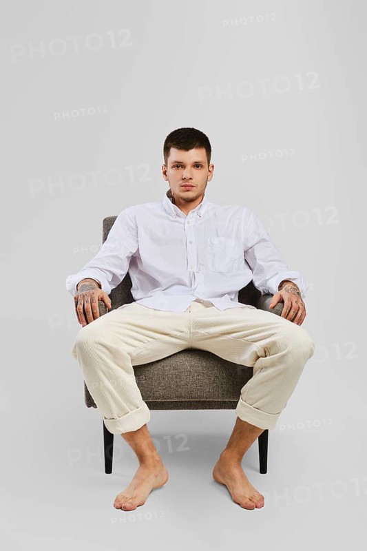 Full length studio portrait of young barefoot man in white shirt and ivory trousers sitting in armchair