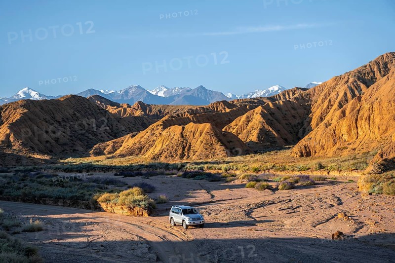 Off-road vehicle driving through a canyon, landscape of eroded hills at sunrise, badlands, white mountain peaks of the Tian Shan Mountains in the background, Canyon of the Forgotten Rivers, Issyk Kul, Kyrgyzstan, Asia