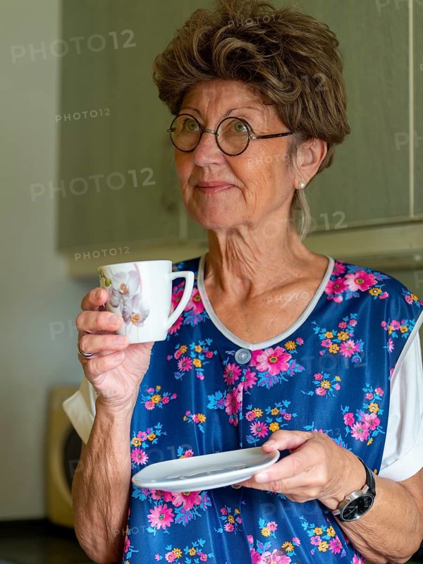 Granny with old smock apron, glasses and wig in the kitchen, holding coffee cup, Germany, Europe