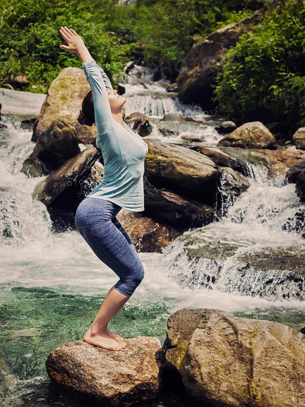 Vintage retro effect hipster style image of sporty fit woman doing yoga asana Utkatasana (chair pose) outdoors at tropical waterfall standing on stone