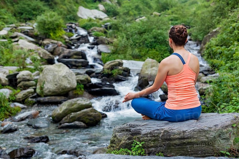 Woman in Hatha yoga asana Padmasana outdoors at tropical waterfall