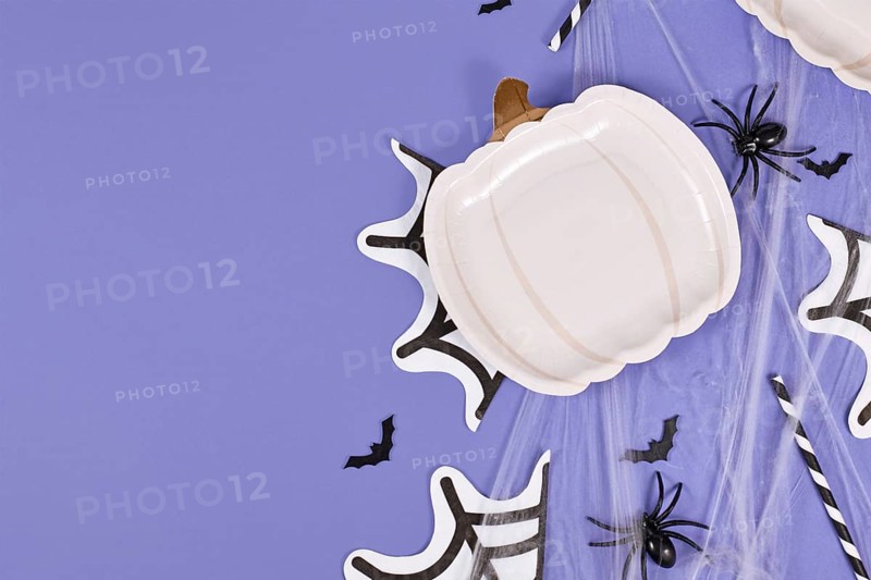 Halloween party flat lay with pumpkin shaped plate, spiders, bats and spiderwebs on violet background with copy space
