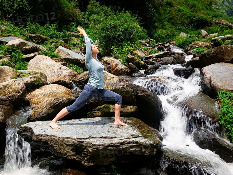 Yoga outdoors, sporty fit woman doing Ashtanga Vinyasa Yoga asana Virabhadrasana 1 Warrior pose posture at waterfall in HImalayas mountains. Vintage retro effect filtered hipster style image