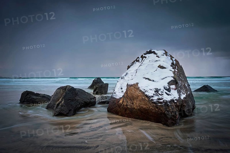 Rocks covered with snow on Norwegian sea beach in fjord in stormy weather with clouds. Skagsanden beach, Flakstad, Lofoten islands, Norway. Long exposure motion blur