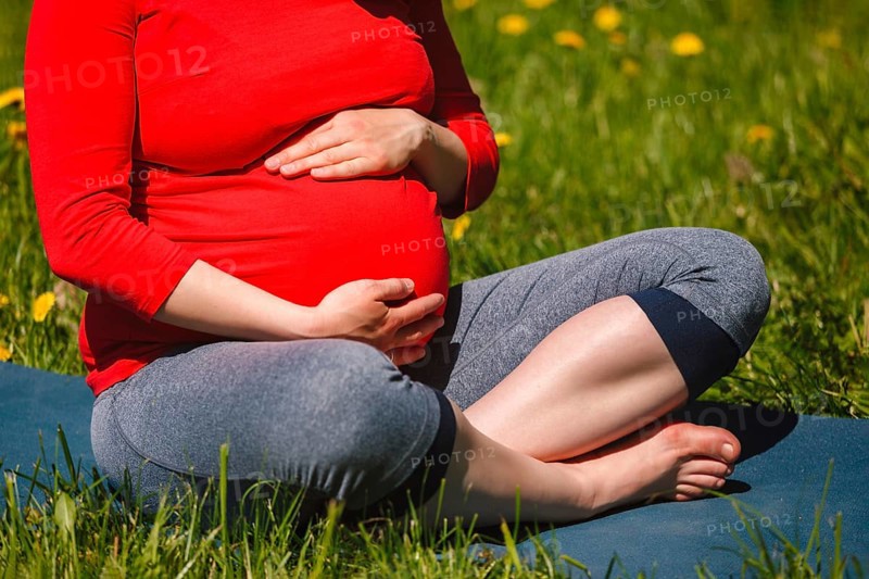 Pregnancy yoga exercise, pregnant woman doing asana Sukhasana easy yoga pose holding her abdomen outdoors on grass lawn with dandelions in summer