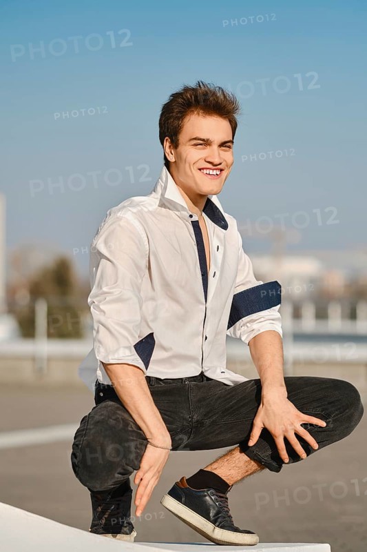 Young man in unfastened white shirt sits on squats on the street