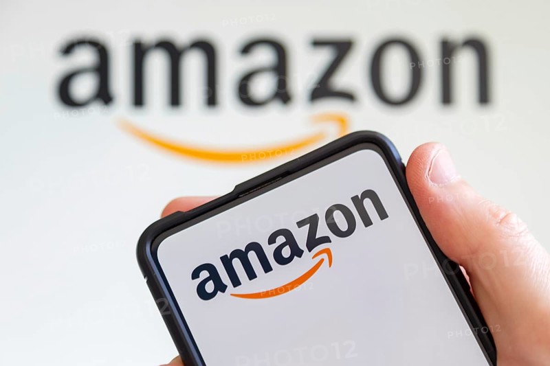 Hand holding mobile phone with Amazon logo online marketplace online trading ecommerce computer screen