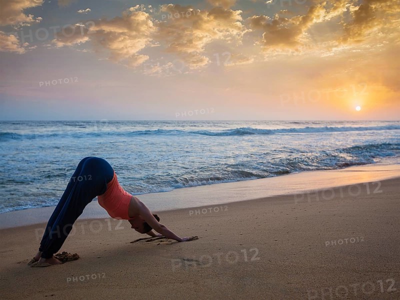 Yoga outdoors, young sporty fit woman doing Ashtanga Vinyasa yoga asana Adho mukha svanasana, downward facing dog, at tropical beach on sunset. Kerala, India, Asia