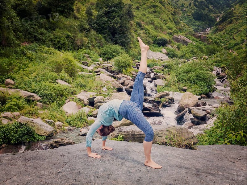 Yoga outdooors, woman doing yoga asana eka pada urdva dhanurasana Upward Bow Pose back benkd outdoors at waterfall in Himalayas. Vintage retro effect filtered hipster style image