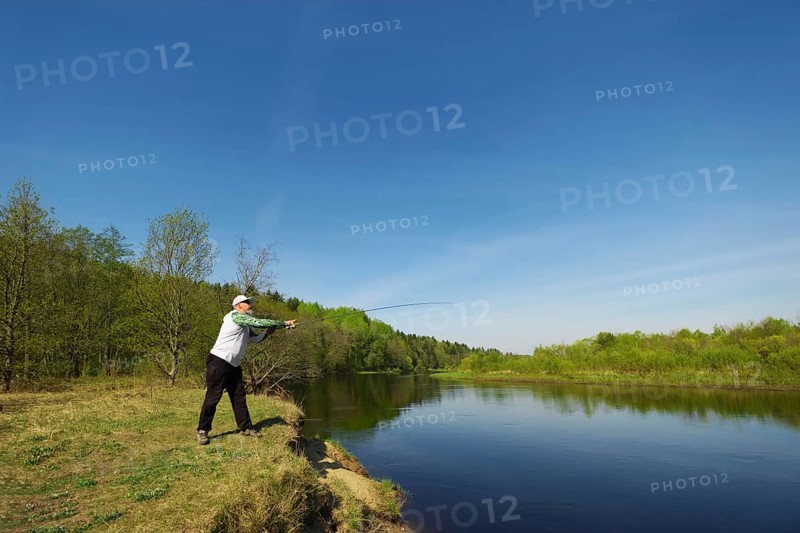 Fisherman with a spinning rod catching fish on a river at sunny summer day with green trees at background. Outdoor weekend activity. Photo with shallow depth of field taken at wide open aperture