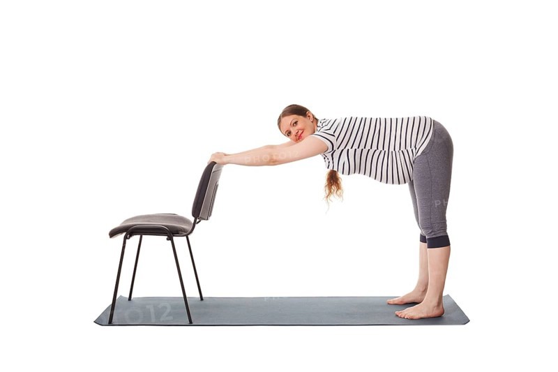 Pregnancy yoga exercise, pregnant woman doing yoga asana Uttanasana Standing Forward Fold Pose with hands on chair easy variation isolated on white background