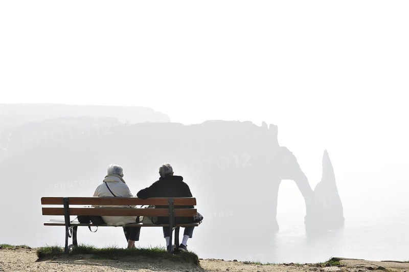 Elderly tourists sitting on bench looking at the Porte D'Aval, a natural arch in the chalk cliffs at Etretat, Côte d'Albâtre, Upper Normandy, France, Europe