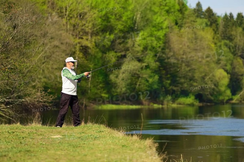 Fisherman with a spinning rod catching fish on a river at sunny summer day with green trees at background. Outdoor weekend activity. Photo with shallow depth of field taken at wide open aperture