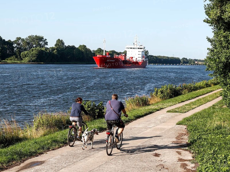 The chemical oil tanker Golfstraum sails on the Kiel Canal, cyclist with dog on cycle path along the canal, Rendsburg, 16 July 2023