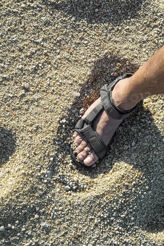 Close-up of man's foot in sandal standing on beach with coarse sea sand grains