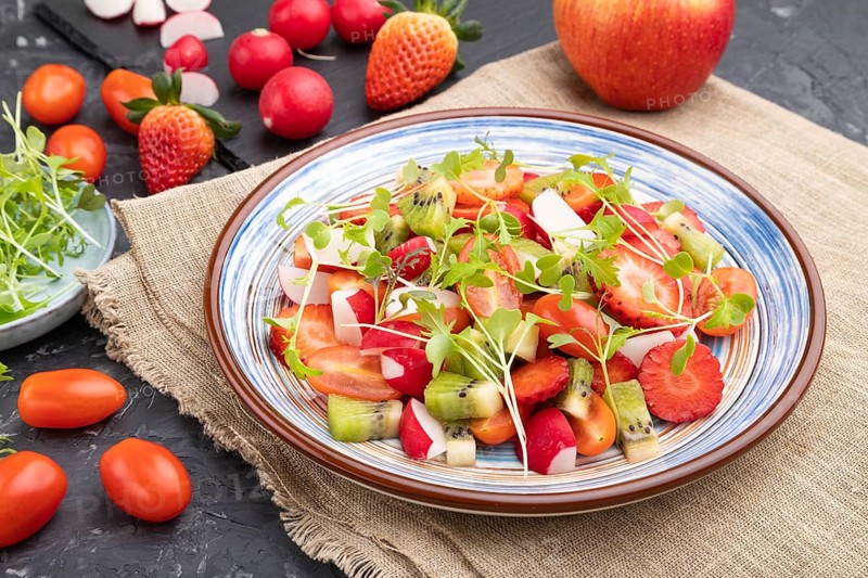 Vegetarian fruits and vegetables salad of strawberry, kiwi, tomatoes, microgreen sprouts on black concrete background and linen textile. Side view, close up