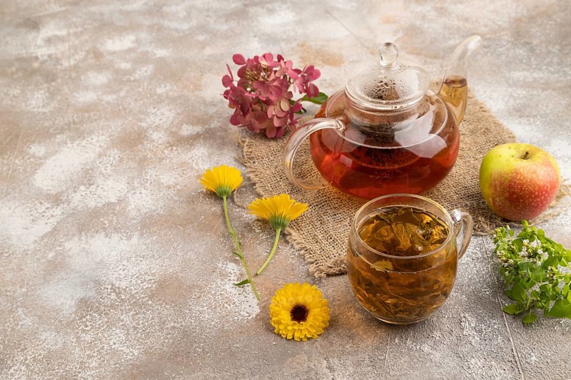 Red tea with herbs in glass teapot on brown concrete background and linen textile. Healthy drink concept. Side view, copy space