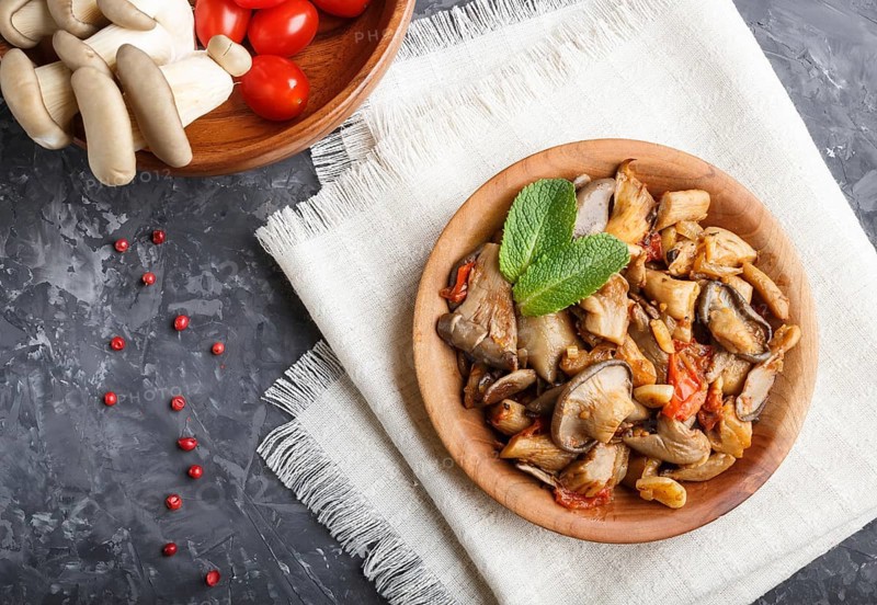 Fried oyster mushrooms with tomatoes in wooden plate on black concrete background. top view, close up