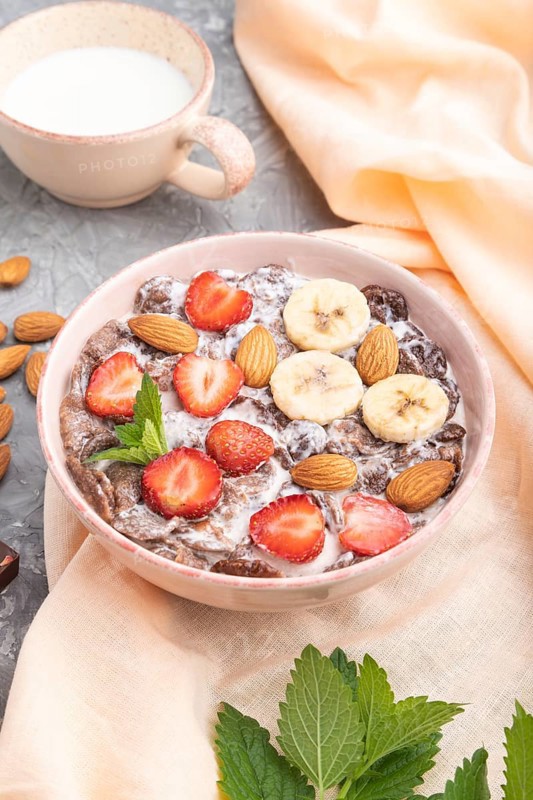 Chocolate cornflakes with milk, strawberry and almonds in ceramic bowl on gray concrete background and orange linen textile. Side view, close up