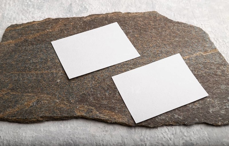 White paper business card, mockup with natural stone on gray concrete background. Blank, side view, still life, copy space