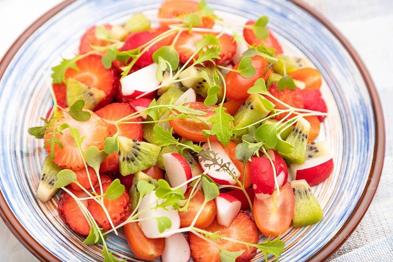 Vegetarian fruits and vegetables salad of strawberry, kiwi, tomatoes, microgreen sprouts on white concrete background and linen textile. Side view, close up, selective focus