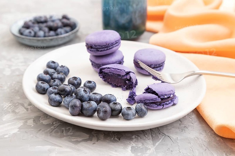 Purple macarons or macaroons cakes with blueberries on white ceramic plate on a gray concrete background and orange textile. Side view, close up, selective focus