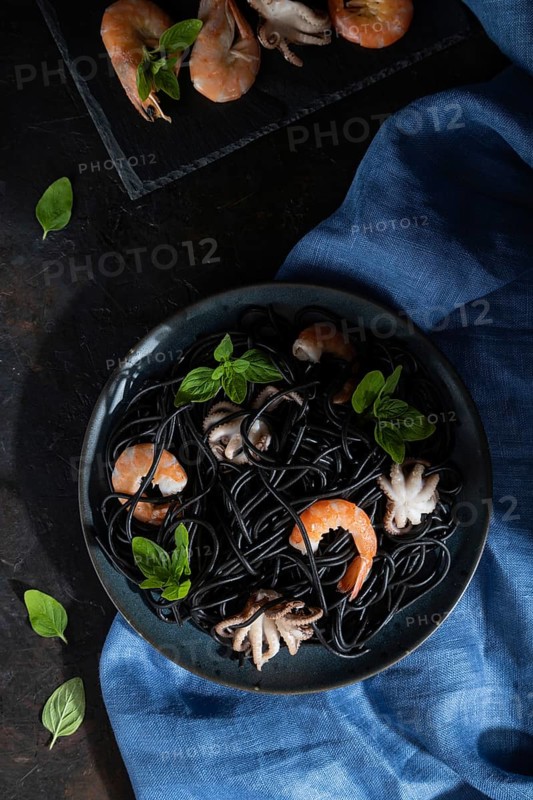 Black cuttlefish ink pasta with shrimps or prawns and small octopuses on black concrete background and blue textile. Low key, hard light. Top view, flat lay, close up