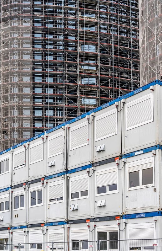 Large construction site, construction of a high-rise residential building, scaffolded shell, already fitted with windows, in front of it containers for offices, accommodation, washrooms, storage, for the construction workers