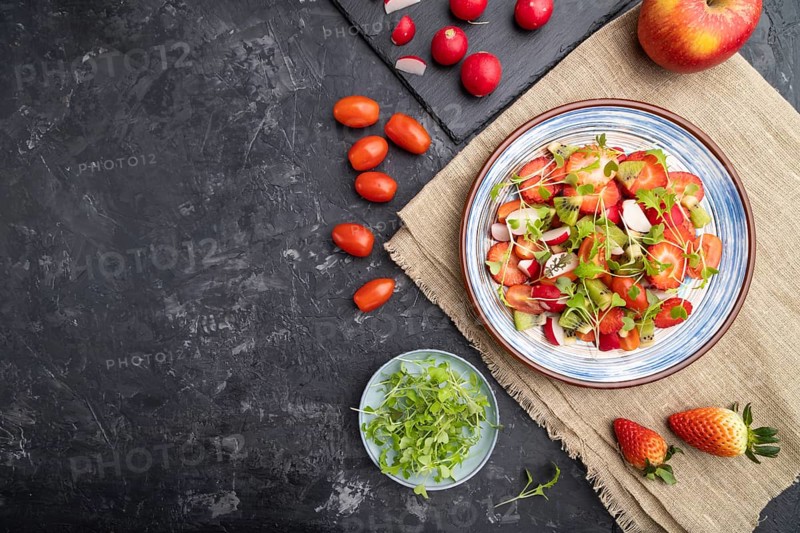 Vegetarian fruits and vegetables salad of strawberry, kiwi, tomatoes, microgreen sprouts on black concrete background and linen textile. Top view, flat lay, copy space