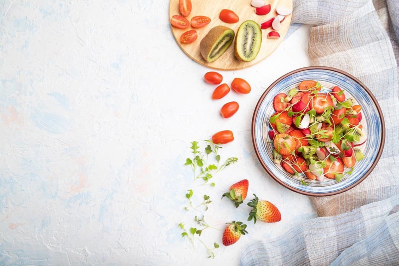 Vegetarian fruits and vegetables salad of strawberry, kiwi, tomatoes, microgreen sprouts on white concrete background and linen textile. Top view, flat lay, copy space