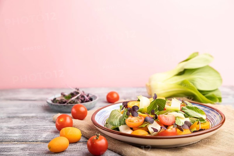 Vegetarian salad of pac choi cabbage, kiwi, tomatoes, kumquat, microgreen sprouts on gray and pink background and linen textile. Side view, close up, selective focus, copy space