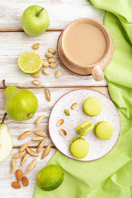 Green macarons or macaroons cakes with cup of coffee on a white wooden background and green linen textile. Top view, flat lay, close up