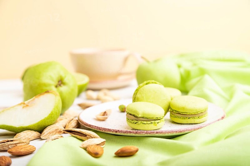 Green macarons or macaroons cakes with cup of coffee on a white wooden background and green linen textile. Side view, close up, selective focus