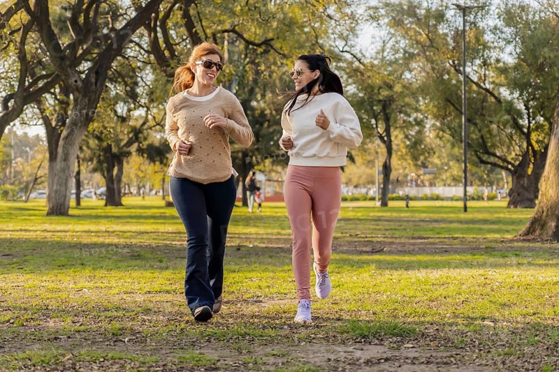 Two women jog side by side in a sunlit park, smiling as they embrace an active lifestyle. Surrounded by lush trees, they enjoy their outdoor exercise and friendship in a serene natural setting