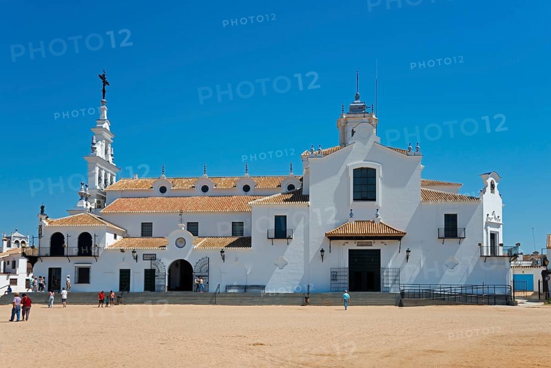Eine weiße Kirche mit typischer spanischer Architektur vor einem klaren blauen Himmel und Sandboden, mit einigen Menschen im Vordergrund, Kirche, Ermita del Rocío, Wallfahrtskirche, Wallfahrtsort, El Rocío, Rocío, Rocio, Huelva, Andalusien, Spanien