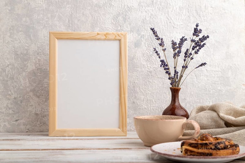 White wooden frame mockup with lavender in ceramic vase, linen textile, cup of coffee and bun on gray concrete background. Blank, vertical orientation, still life