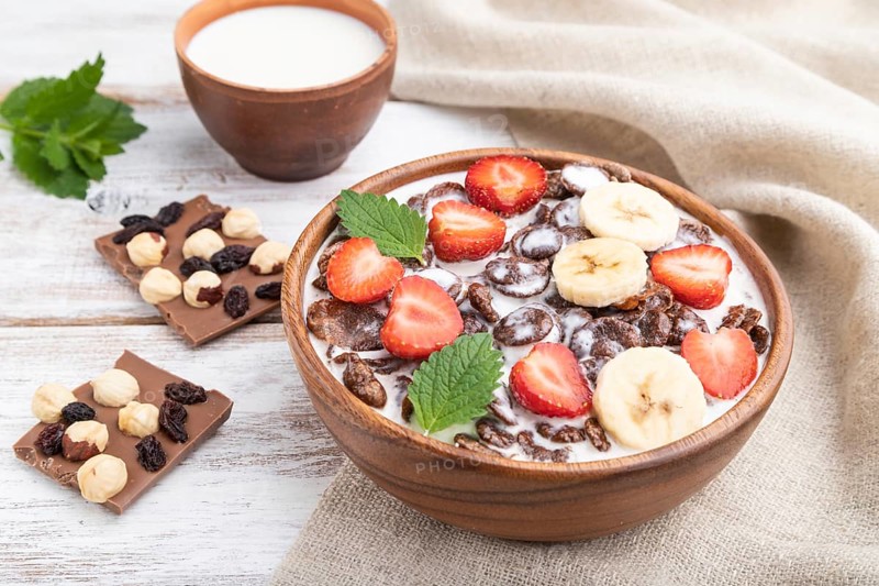 Chocolate cornflakes with milk and strawberry in wooden bowl on white wooden background and linen textile. Side view, close up