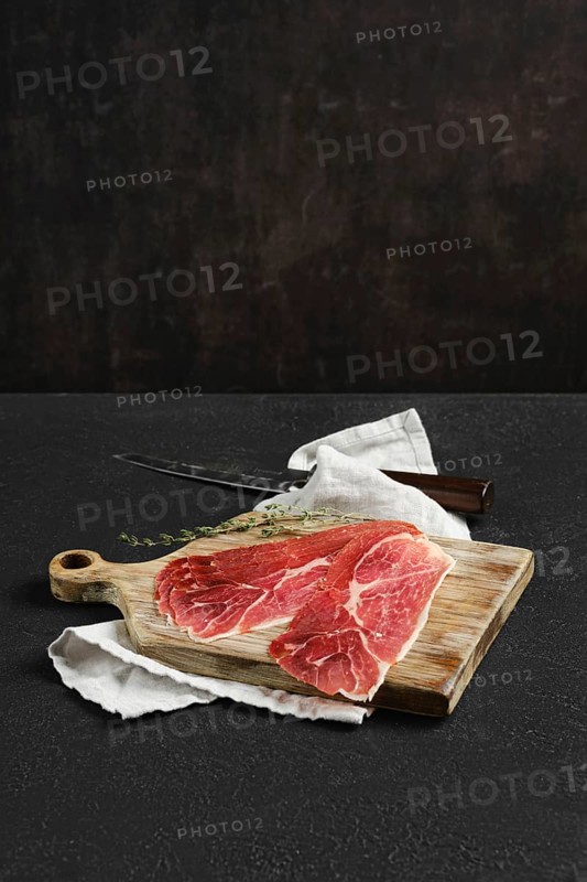 Thin slices of jamon iberico on a wooden cutting board, accompanied by a knife and a sprig of thyme. A white cloth adds a rustic touch to the dark, textured background, enhancing the presentation.