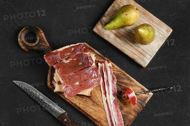 Cured duck meat on a wooden board next to two ripe pears. A knife rests nearby, highlighting the preparation of a gourmet meal in a modern kitchen.