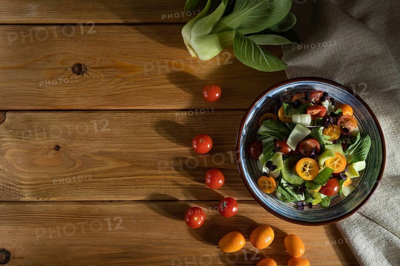 Vegetarian salad of pac choi cabbage, kiwi, tomatoes, kumquat, microgreen sprouts on a wooden background and linen textile. Hard light, contrast. Top view, flat lay, copy space