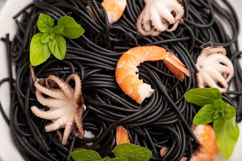 Black cuttlefish ink pasta with shrimps or prawns and small octopuses on gray wooden background and blue textile. Top view, close up, selective focus