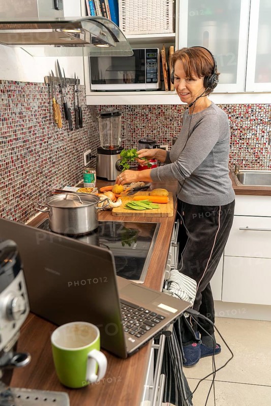 Woman, mid-50s, works from home, with laptop and communicates with colleagues via headset, home office, in the kitchen, while cooking